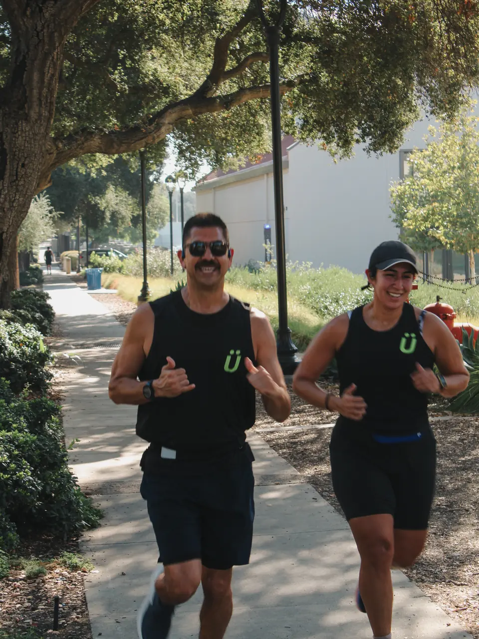 Two runners in green Ü logo tanks smiling side by side on a tree lined path